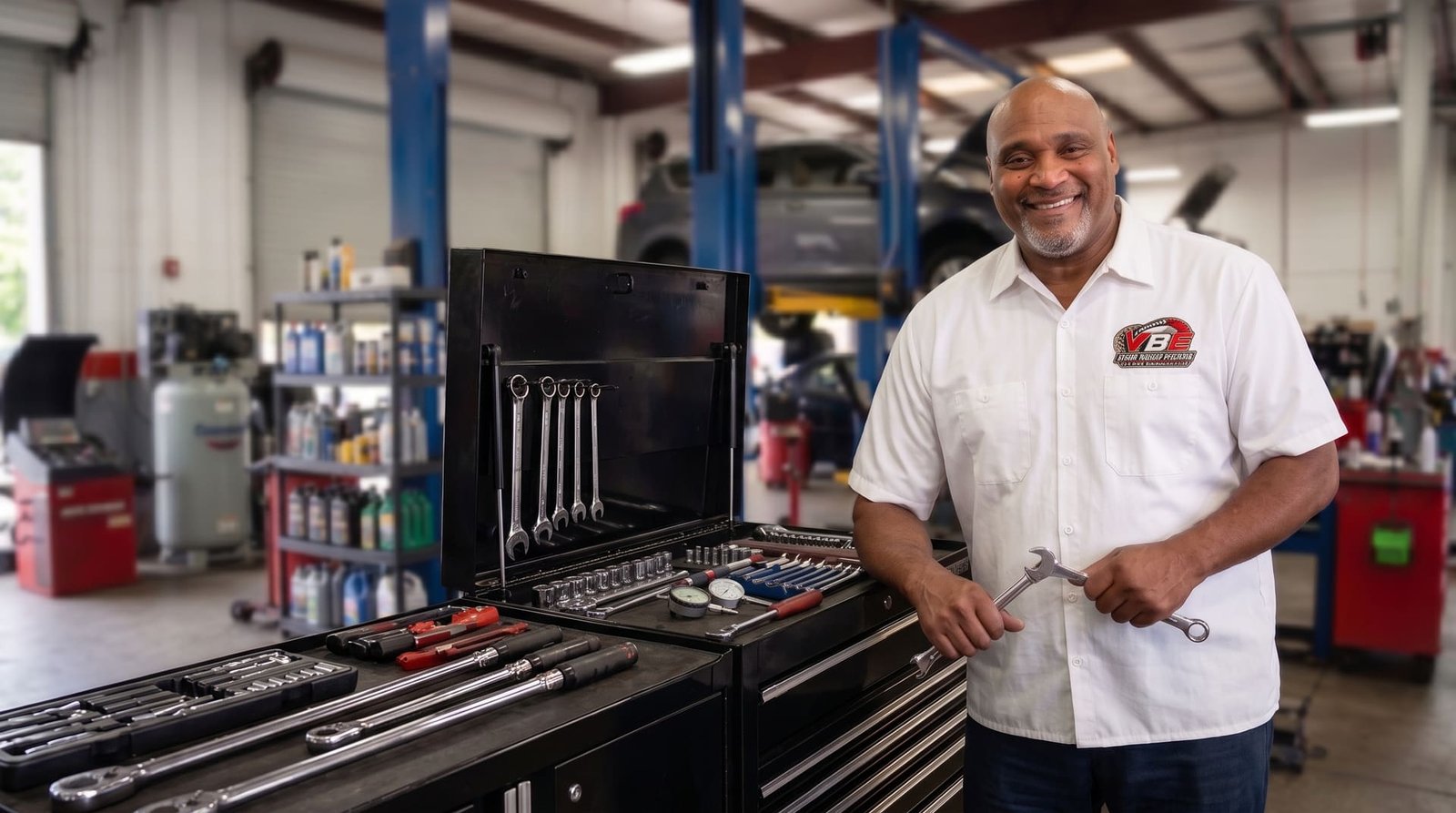 Inside YBE Auto Repair Center: an open tool chest and a technician holding a wrench, with a vehicle up on the lift behind him
